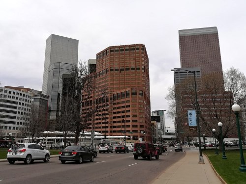 Cityscape featuring modern skyscrapers, a busy street with cars, and trees along the sidewalk under a cloudy sky.
