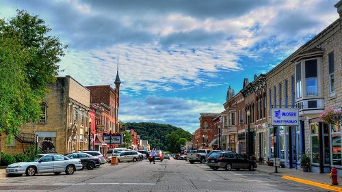 A charming small-town street lined with shops, cars, and a cloudy sky in the background.