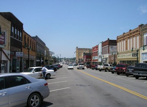 A view of a downtown street lined with shops and parked cars under a clear blue sky.