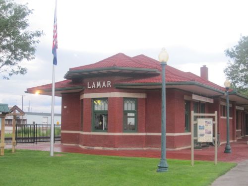 Historic Lamar train station with red brick exterior, green windows, and a flagpole in front.