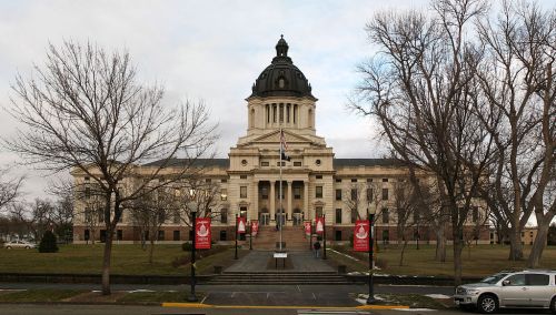 Historic building with a dome, flanked by trees and banners, set against a cloudy sky.