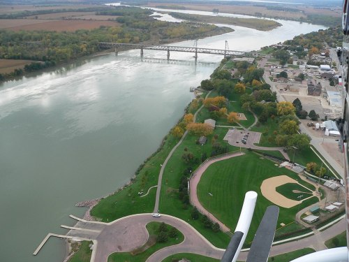 Aerial view of a river, bridge, and a park with a baseball field along the shoreline, surrounded by trees and buildings.