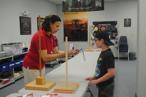 A woman demonstrates a science experiment to a boy at a table in a classroom setting.