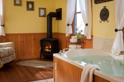 Cozy bathroom with a soaking tub, a wood stove, and a comfortable chair, featuring warm yellow walls and natural light.