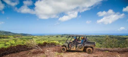Two people in an off-road vehicle on a scenic hillside, overlooking lush greenery and the ocean under a blue sky.