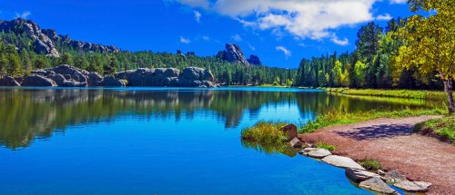 A serene lake surrounded by lush trees and rocky formations under a bright blue sky with fluffy clouds.
