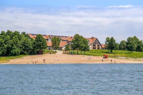 A sandy beach by a lake with people enjoying the sun, surrounded by green trees and a large building in the background.