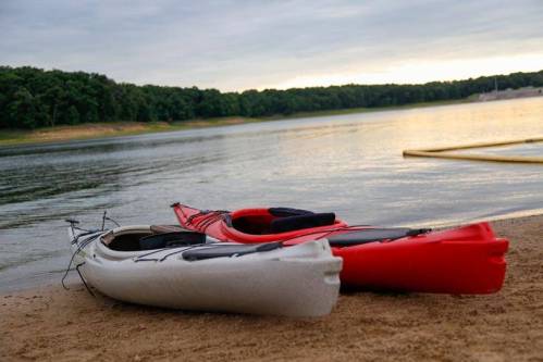Two kayaks, one white and one red, resting on a sandy beach by a calm lake surrounded by trees.