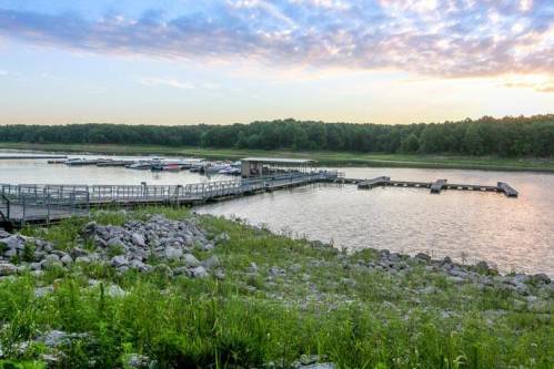 A serene lake at sunset with a wooden dock and boats, surrounded by lush greenery and rocky shoreline.