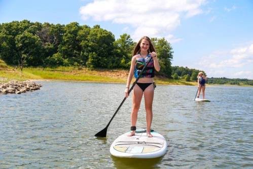 A girl stands on a paddleboard in a lake, smiling and holding a paddle, with trees and blue sky in the background.