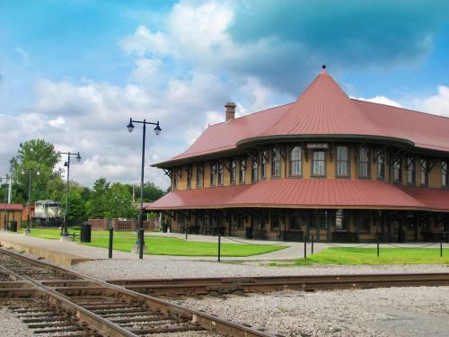 Historic train station with a red roof, surrounded by green grass and a cloudy blue sky, near railway tracks.