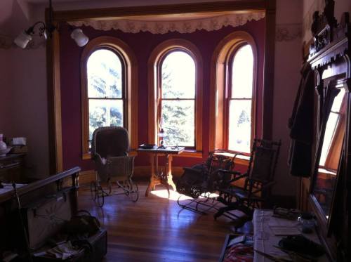 A cozy room with three large windows, a wooden table, and vintage chairs, illuminated by natural light.