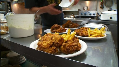 Plates of fried chicken and fries on a kitchen counter, with a chef preparing food in the background.