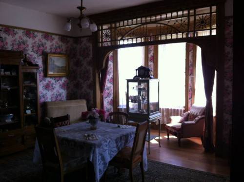 A vintage dining room with floral wallpaper, a wooden table, and a display cabinet, featuring a sunny window nook.