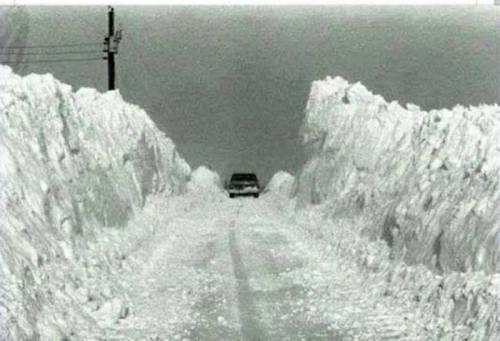 A car drives down a snow-covered road, flanked by tall snowbanks on either side.