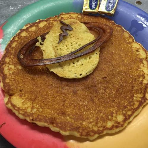 A large pancake topped with a smaller pancake shaped like Saturn, served on a colorful plate with butter packets.