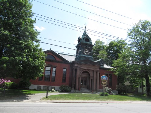 Historic brick library building with a clock tower, surrounded by trees and a clear blue sky.