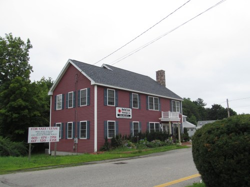 A two-story red house with a gray roof, featuring a "For Sale/Lease" sign in front and surrounded by greenery.