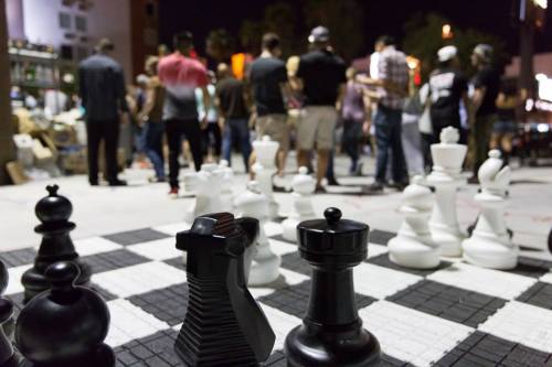 A large outdoor chess game at night, with people gathered around, focusing on the black and white pieces in the foreground.