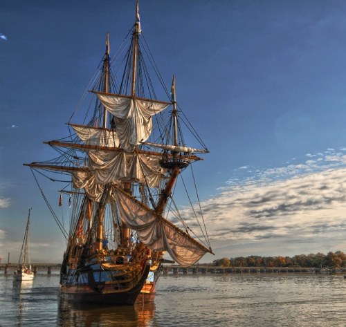 A tall ship with billowing sails docked on calm waters under a clear blue sky.