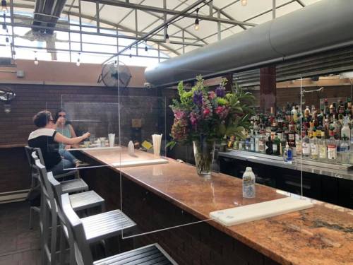 A bar with a clear divider, featuring a vase of flowers and a well-stocked liquor shelf in the background.