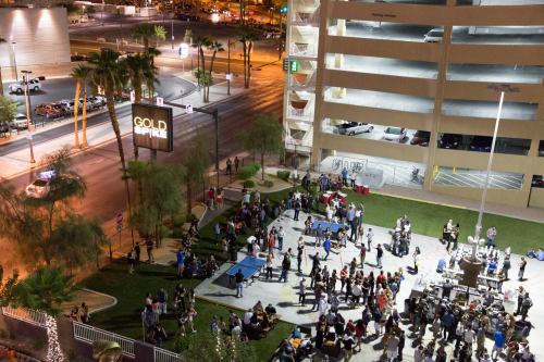 Aerial view of a lively outdoor gathering at night near a parking garage, with people socializing and city lights in the background.
