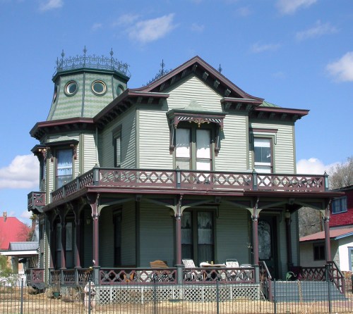 Victorian-style house with a turret, intricate woodwork, and a wraparound porch against a blue sky.