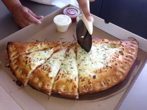 A hand using a pizza cutter to slice a large calzone in a pizza box, with dipping sauces nearby.