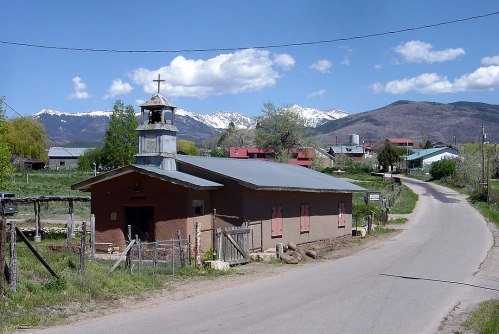A small church with a bell tower beside a rural road, surrounded by mountains and greenery under a blue sky.