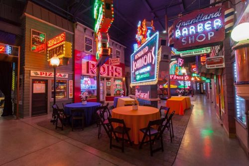 Colorful vintage neon signs illuminate a lively indoor street scene with tables and chairs set up for dining.