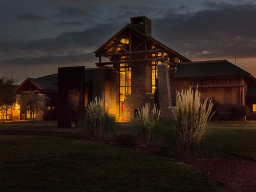 A modern building illuminated at night, surrounded by grass and shrubs under a cloudy sky.
