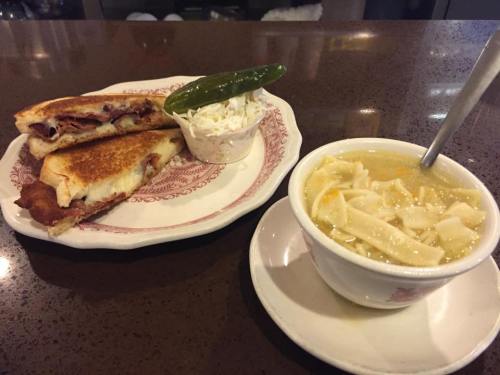 A plate with a grilled sandwich, coleslaw, a jalapeño, and a bowl of chicken noodle soup on a dark countertop.