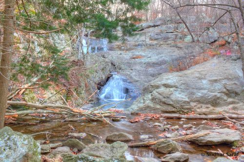 A serene waterfall cascades over rocky terrain, surrounded by trees and autumn foliage.