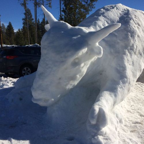 A large snow sculpture of a goat, with detailed features, set against a snowy landscape and blue sky.