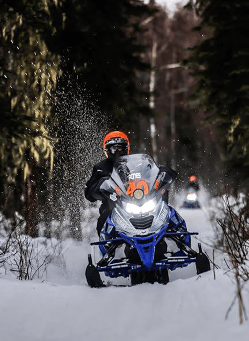 A snowmobiler speeds through a snowy forest trail, kicking up snow, with another snowmobile in the background.