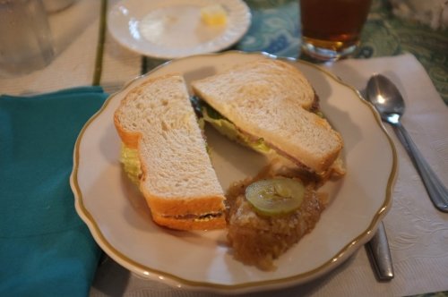 A plate with a sandwich cut in half, accompanied by a small serving of dessert and a drink in the background.
