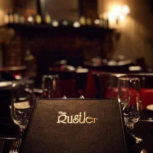 A dimly lit restaurant table set with glasses, featuring a menu titled "The Rustler" in the foreground.