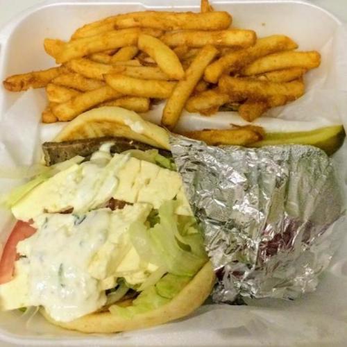 A takeout container with a sandwich filled with lettuce, tomato, and sauce, alongside a serving of crispy French fries.