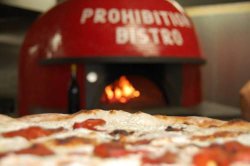 A close-up of a pizza in front of a red oven with "Prohibition Bistro" written on it, flames visible inside.