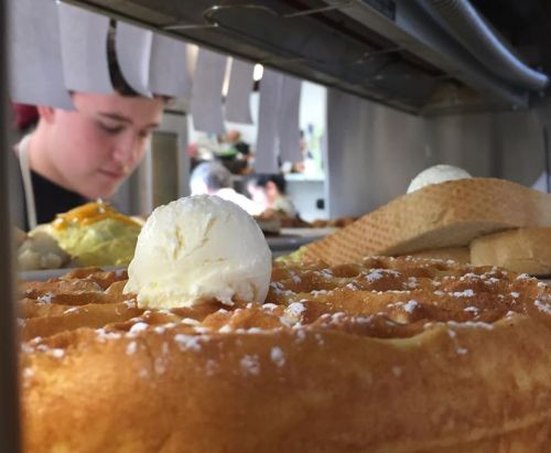 A close-up of a waffle topped with a scoop of ice cream, with a person preparing food in the background.