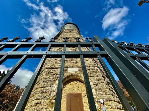 A stone tower viewed from below, surrounded by a black metal fence against a blue sky with clouds.