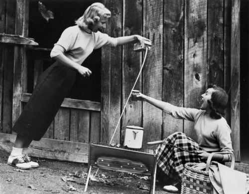 Two women interact outdoors, one pouring from a container while the other reaches out, with a rustic wooden backdrop.