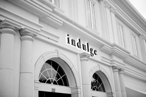A black and white photo of a building facade featuring the word "indulge" above an arched entrance with columns.