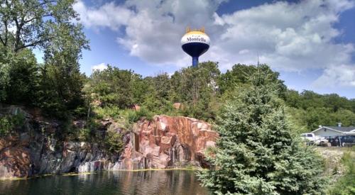 A scenic view of a water tower labeled "Monticello" above a rocky landscape and a calm pond under a blue sky.