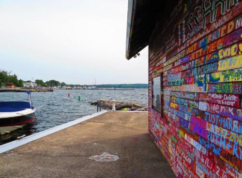 Colorful graffiti covers a brick wall near a dock, with a boat and calm water in the background.