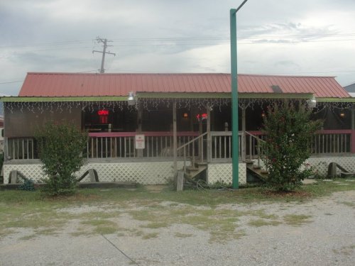 A small restaurant with a red metal roof, porch, and "Open" sign, surrounded by greenery and gravel parking.