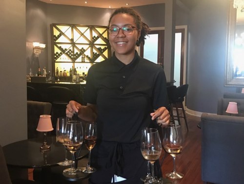 A smiling woman in a black shirt stands behind a bar with wine glasses in front of her.