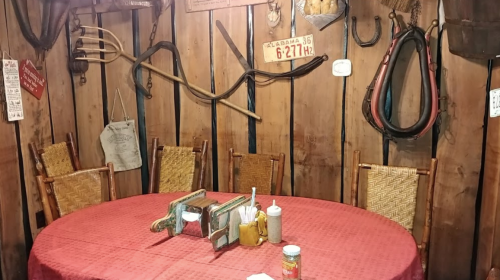 A rustic dining area with a round table, wicker chairs, and vintage farm tools on wooden walls.