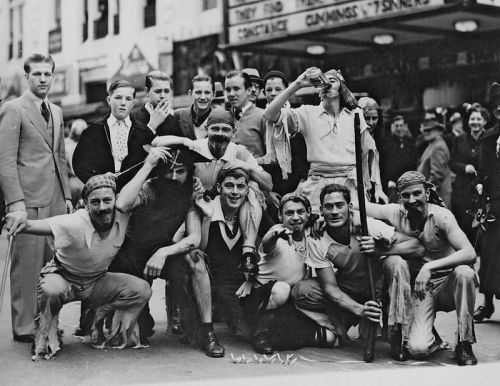 A group of men in vintage clothing posing playfully, some in costumes, in a lively street scene.