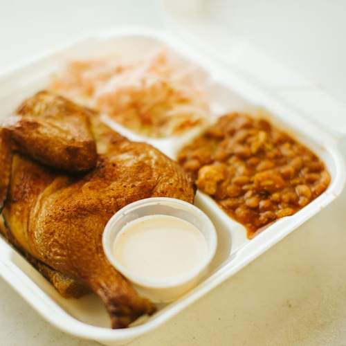 A takeout container with seasoned chicken, baked beans, coleslaw, and a small cup of sauce.